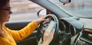 Driver holding a smartphone while operating a vehicle, with one hand on the steering wheel and eyes directed toward the phone