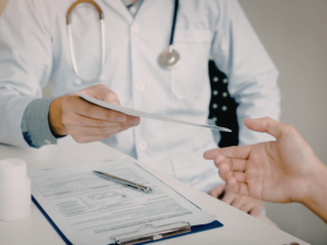Doctor handing a patient a medical document across a desk with forms and a stethoscope