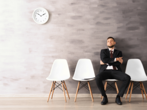 Man in a suit waits alone on a row of chairs under a clock, possibly for a job interview