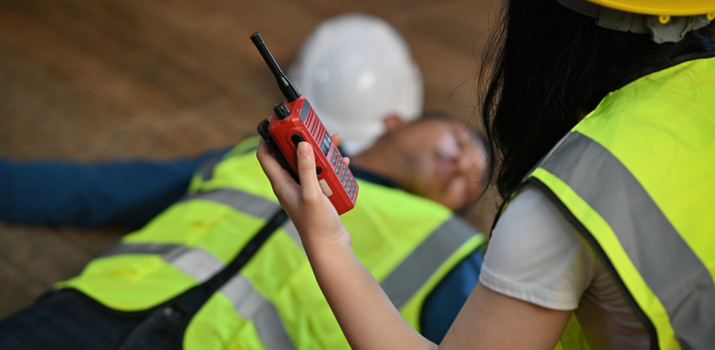 Construction worker using radio beside injured colleague on ground, highlighting workplace accident response