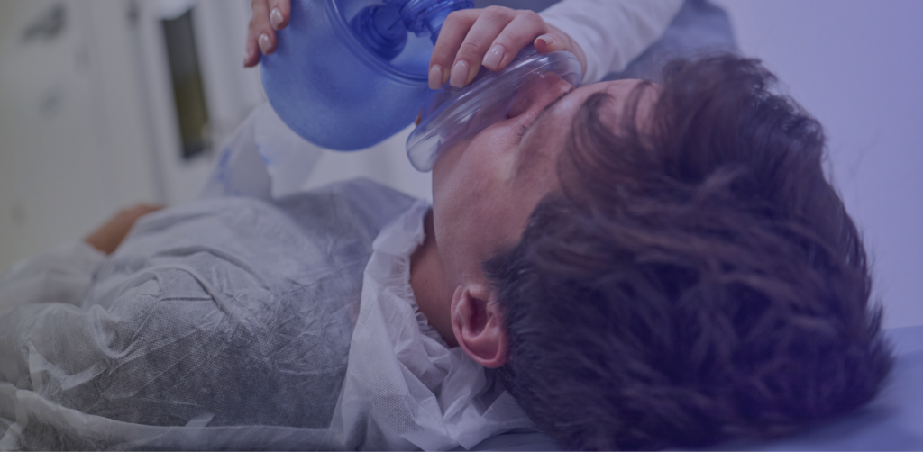 Medical professional using a manual resuscitation bag and oxygen mask to assist a patient lying on a hospital bed