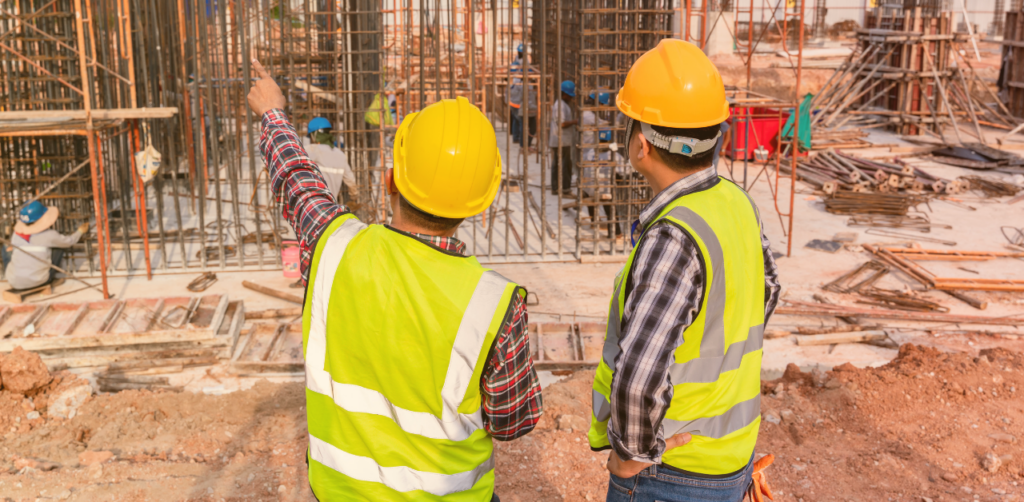 Two construction workers wearing hard hats and reflective vests observing and discussing activity at an active construction site