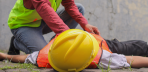 Worker in a safety vest assisting an injured construction worker lying on the ground while wearing a hard hat and reflective gear