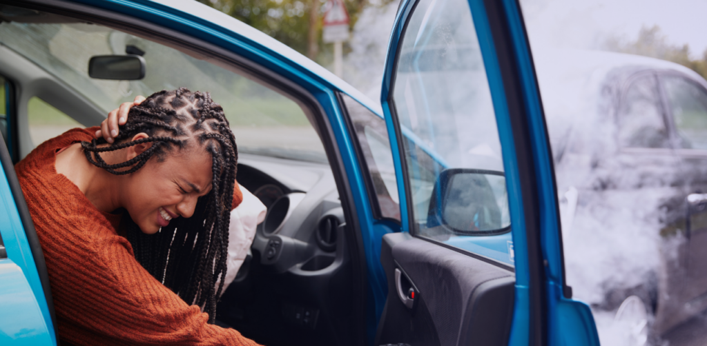 Injured driver sits in an open car door holding their head after a crash, with smoke from another vehicle nearby